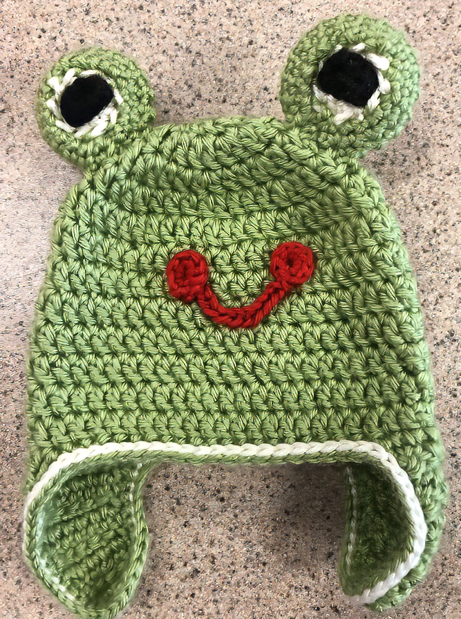 Knit green hat with ear flaps, resembling a frog with big eyes and a red smile.