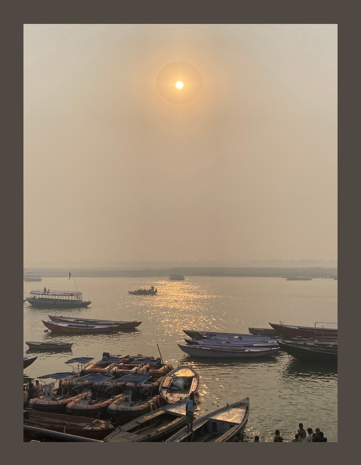 Sunset over a hazy river with reflections shimmering on the water. Numerous boats and a few people gather at the riverside.