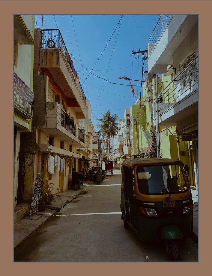 Narrow urban street lined with colorful buildings, laundry hanging from balconies, a parked auto rickshaw, and a distant palm tree under clear blue skies.