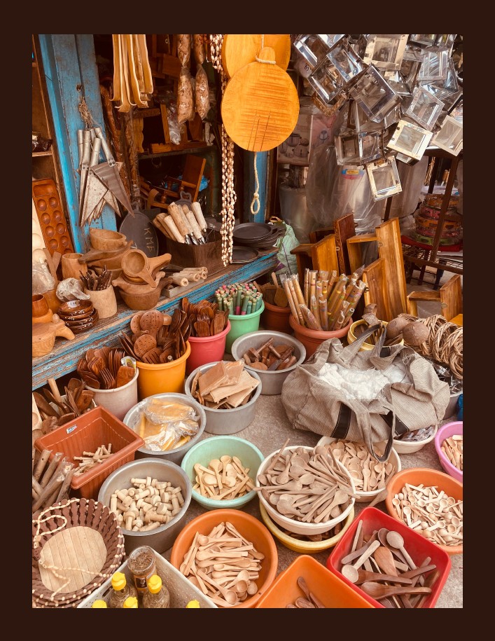 Colorful buckets filled with handcrafted wooden spoons, utensils, and other items are displayed in a rustic market stall.