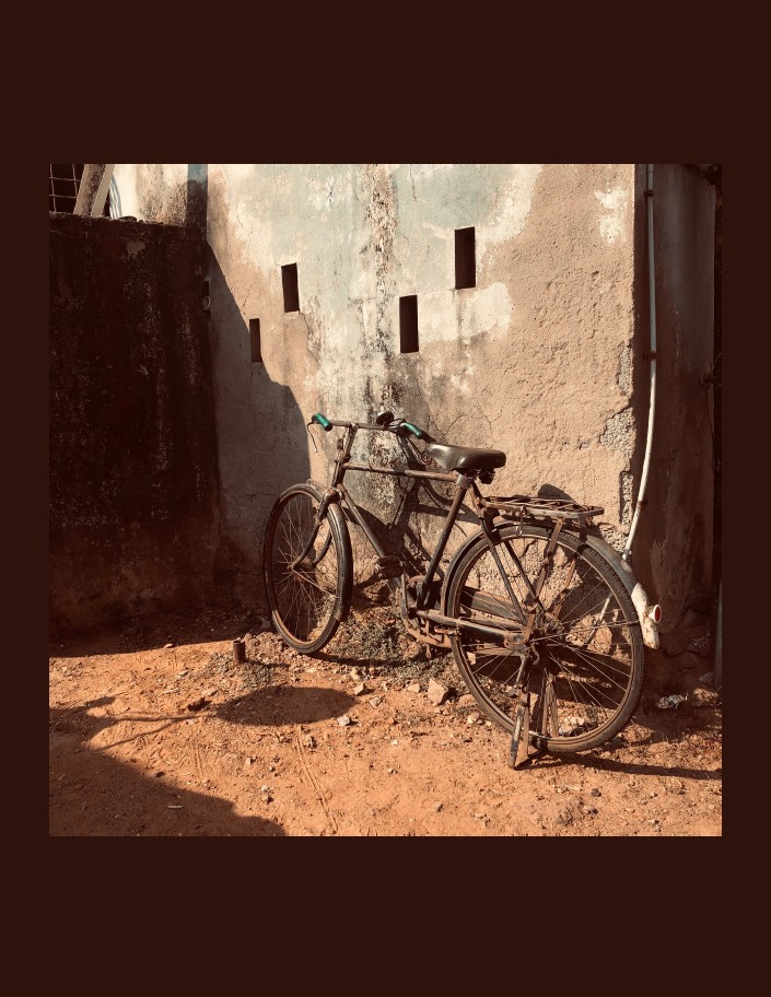 A vintage bicycle with rust leans against an old, textured wall in warm sunlight, casting distinct shadows on the dusty ground.