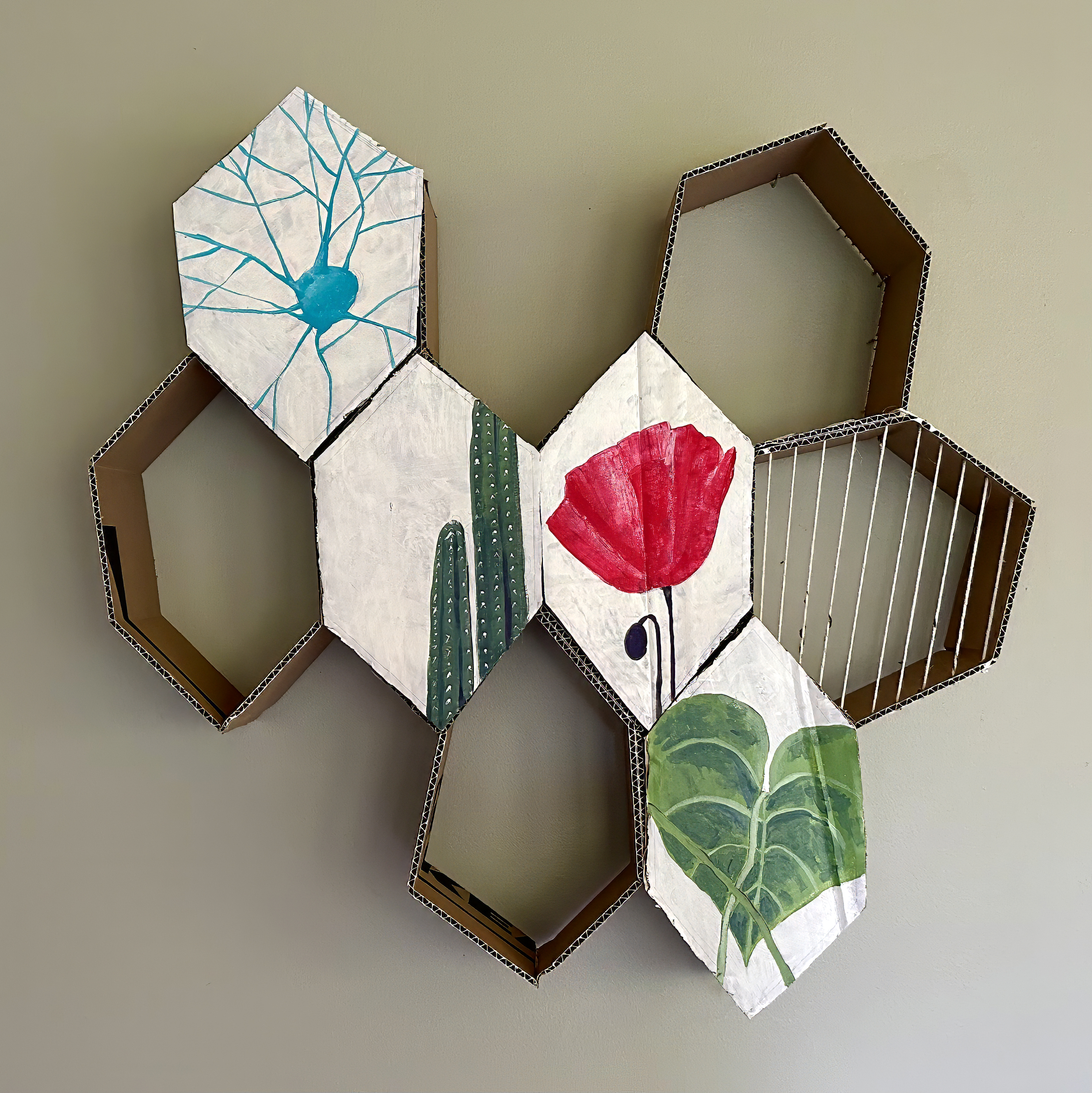 Hexagonal/honeycomb cardboard art piece on a wall, featuring a vibrant red flower, cactus, green leaves, blue abstract design, and one section with strings.