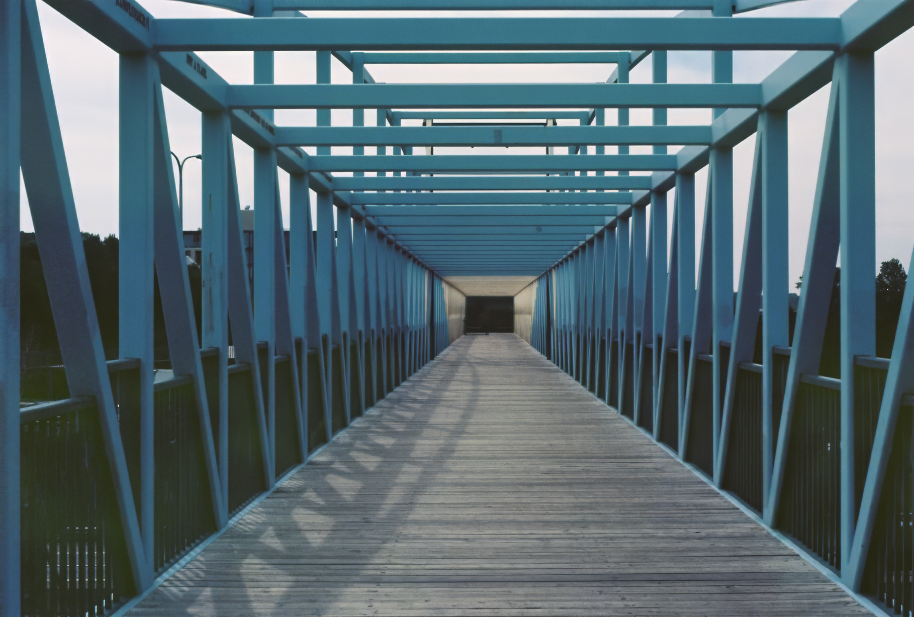 Sky-blue metal pedestrian bridge with geometric patterns and wooden floor forms a tunnel, creating a sense of depth and symmetry on an overcast day.