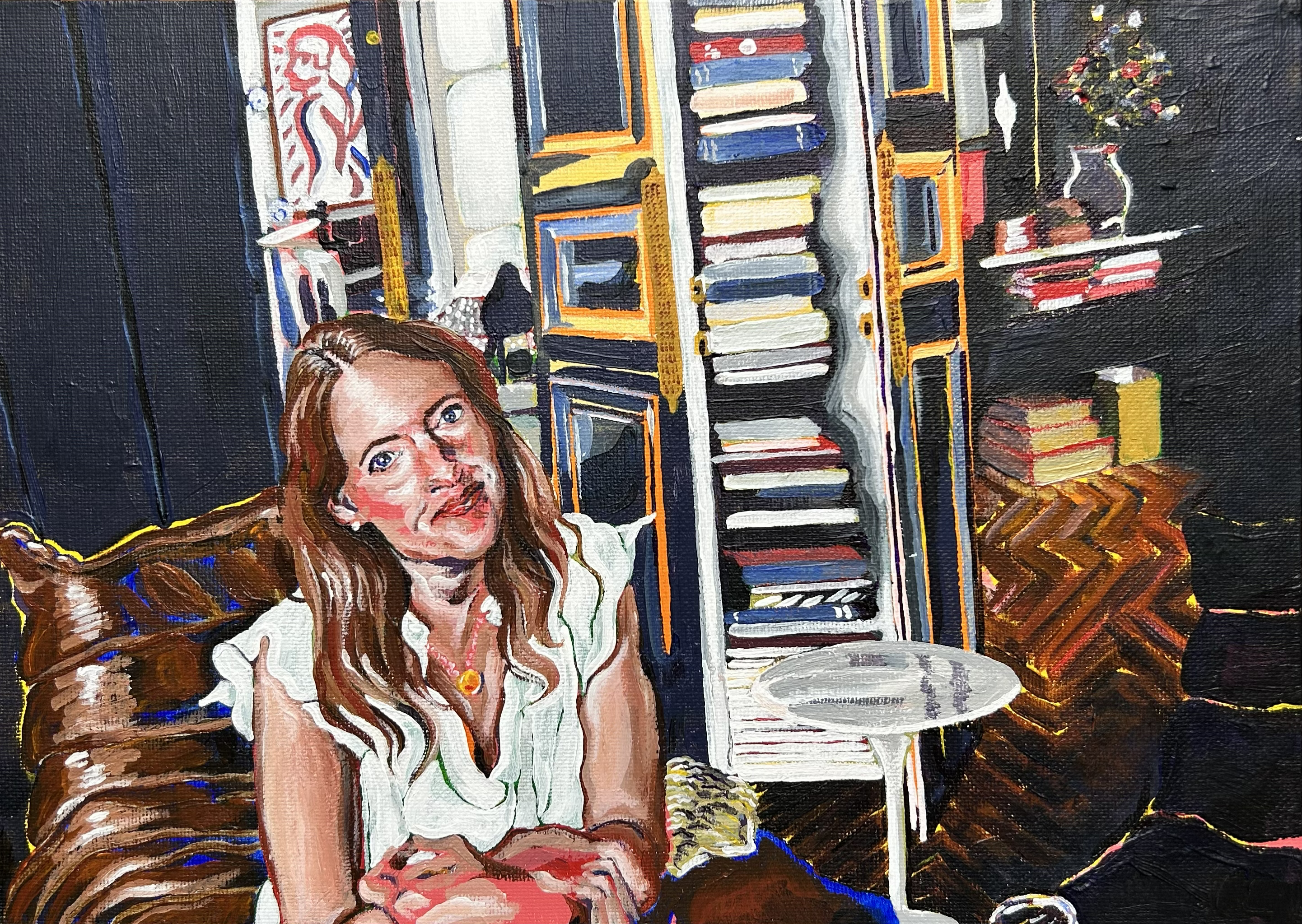 A woman with long brunette hair in a white blouse sits on a brown sofa in a living room, looking at the viewer. Behind her is a tall, colorful stack of books between black shelving.