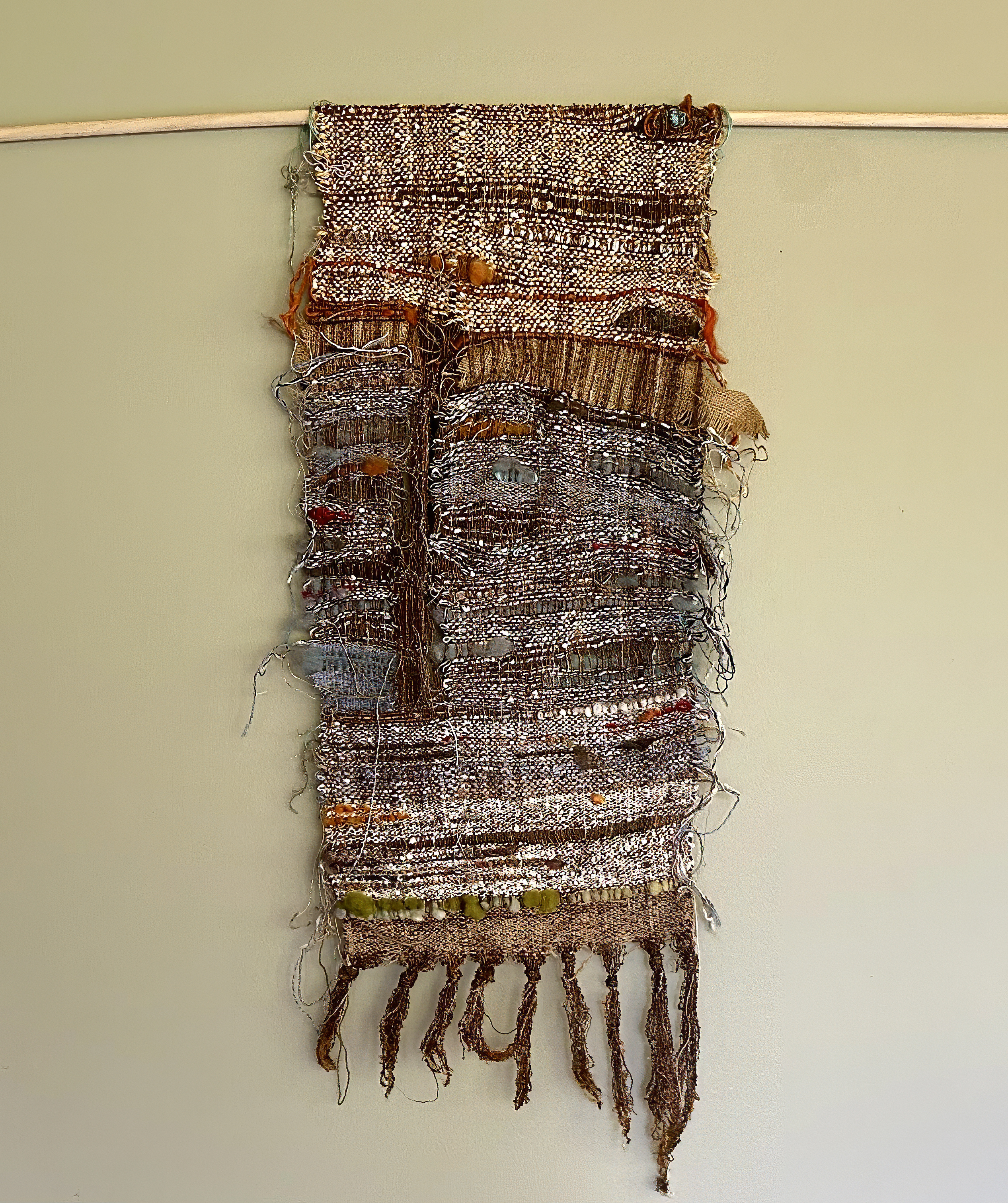 A rustic, handwoven tapestry with earthy tones hangs from a wooden rod against a beige wall. The fabric features textured, frayed edges, and beads.