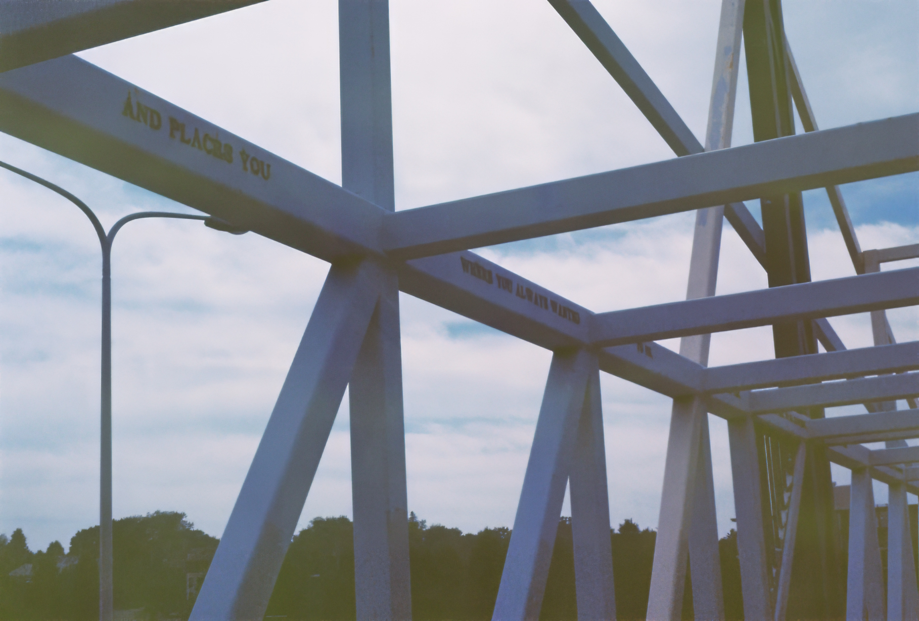 View of a bridge's metal framework against a cloudy sky, with partial text visible on the beams. Trees are seen in the distance below.